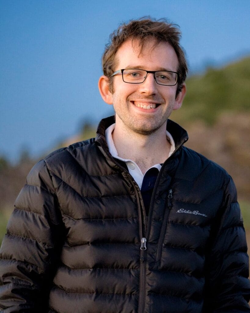 A smiling individual wearing glasses and a black Eddie Bauer puffer jacket stands outdoors with hills and a clear sky in the background.