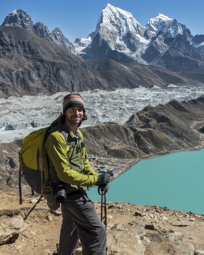 A hiker wearing a green jacket, beanie, and backpack stands on a rocky hill overlooking a turquoise lake and snowy mountain peaks in the background. He holds trekking poles and a camera is hanging from his neck. The sky is clear with abundant sunshine.