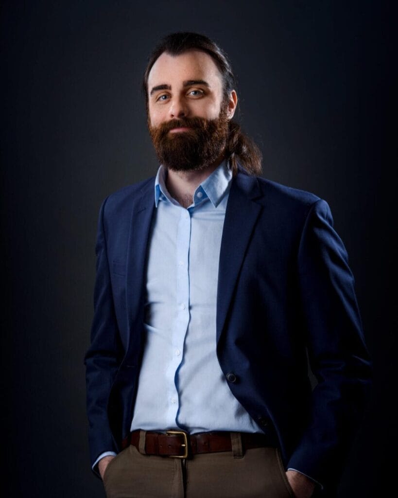 A man with a beard and long hair tied back stands against a dark background. He is wearing a dark blazer over a light blue dress shirt, with his hands in his pants pockets. He has a relaxed and confident expression on his face.