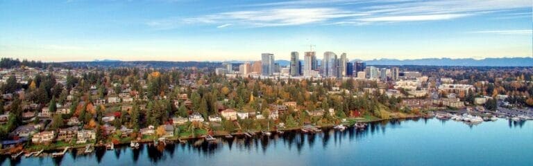professional real estate photography. A panoramic aerial view of Bellevue featuring a mix of high-rise buildings and low-rise residential homes surrounded by trees, all situated along the edge of a calm body of water. The sky above is clear with scattered clouds.
