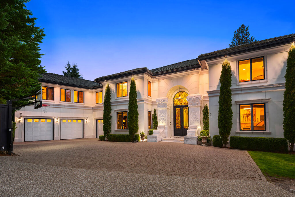 A large modern mansion with white exterior walls, tall windows, and a grand entrance featuring a double door framed by ornate columns. The house has multiple lit-up windows and a three-car garage on the left, surrounded by manicured greenery and a paved driveway.