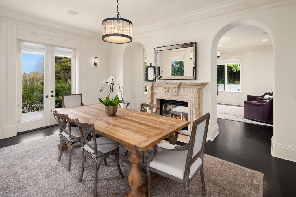 A dining room with a wooden table and six upholstered chairs. A potted orchid is in the center of the table. The room has a chandelier, a fireplace with a mirror above it, and arched doorways. French doors open to a garden, and a living room is visible in the background.