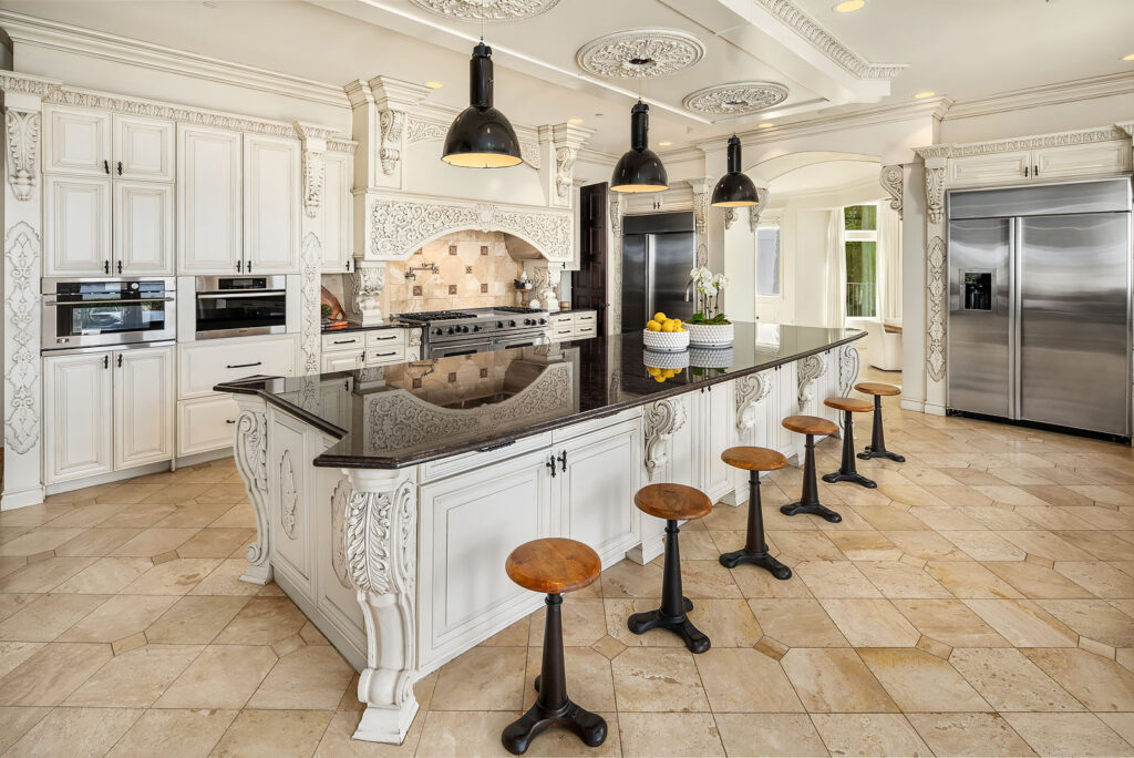 A spacious, elegant kitchen featuring a large, ornate white island with a glossy black countertop and five wooden stools. The background showcases intricate white cabinetry, stainless steel appliances including double ovens, and pendant lighting above the island.