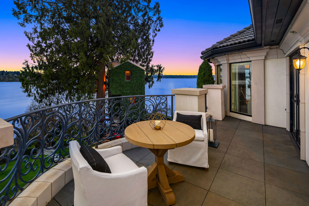 A cozy balcony features cushioned chairs and a wooden round table atop a tiled floor. Ornate iron railings enclose the space, which offers a scenic view of a lake and distant trees under a clear twilight sky. A warm lantern light glows on the right wall.