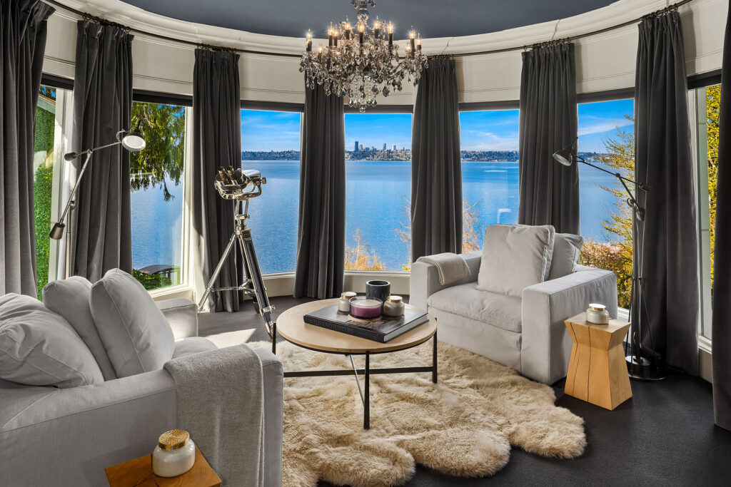 A luxurious living room with large windows offering a panoramic view of a lake and city skyline. The room features plush gray chairs, a furry white rug, a coffee table with candles, a chandelier, and a telescope aimed at the scenery outside.