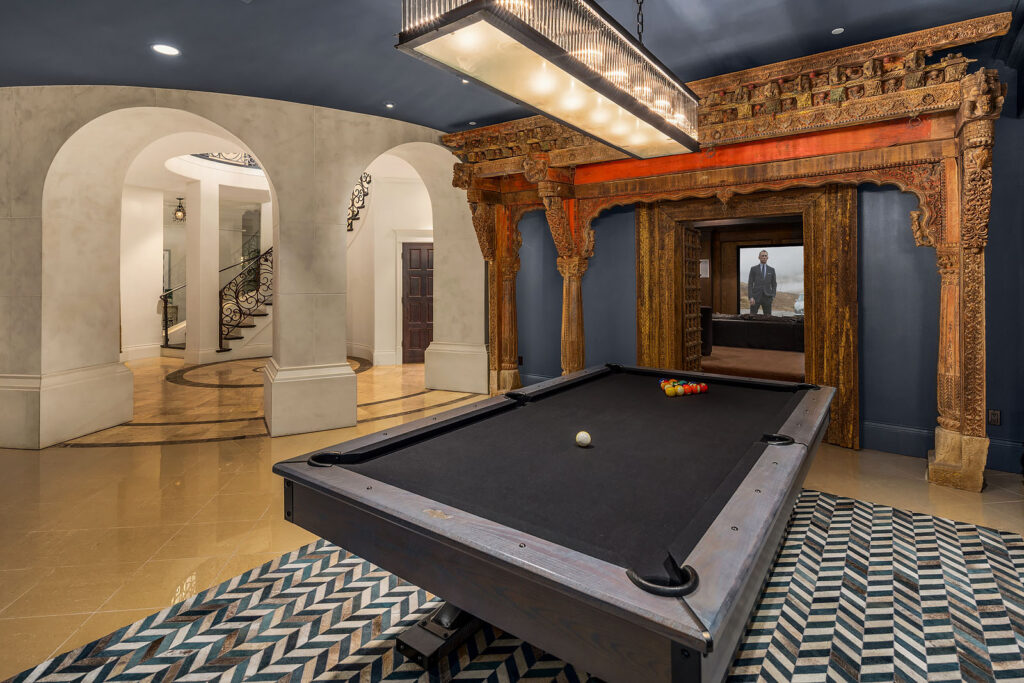 A well-lit game room featuring a pool table with a black felt surface. The room has blue walls, an ornate wooden frame, and a geometric patterned rug. Arched doorways and a grand staircase with intricate railings are visible in the background.