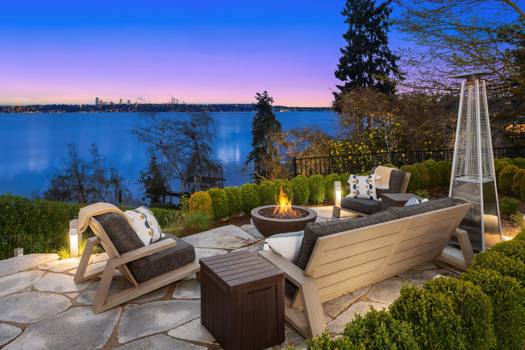 Outdoor patio with comfortable chairs surrounding a fire pit overlooking a serene lake at sunset. The vibrant twilight sky and distant cityscape create a tranquil ambiance. Trees and shrubs border the stone patio, enhancing the natural beauty of the scene.