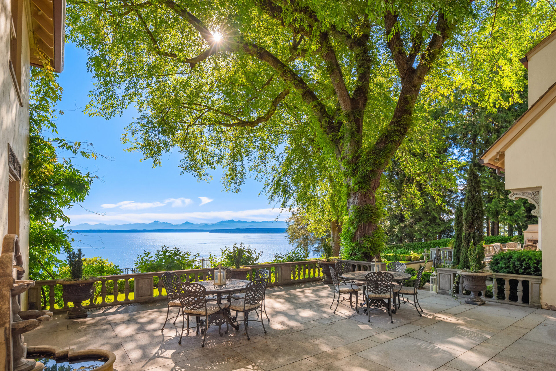 A serene patio with wrought iron furniture is shaded by a large, leafy tree. Beyond the stone balustrade of this Tim Van Asselt Edwin Ivey estate, a picturesque view of a tranquil lake and distant mountains is visible under a bright blue sky. Sunlight filters through the tree branches, casting dappled light.