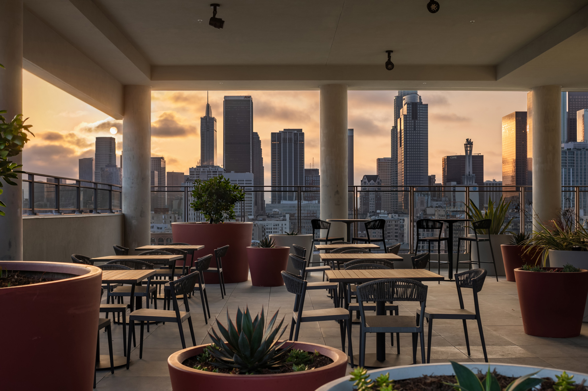 Rooftop terrace with modern seating and planters, offering a stunning view of the Los Angeles skyline at sunset. Ideal for gatherings in an urban setting.