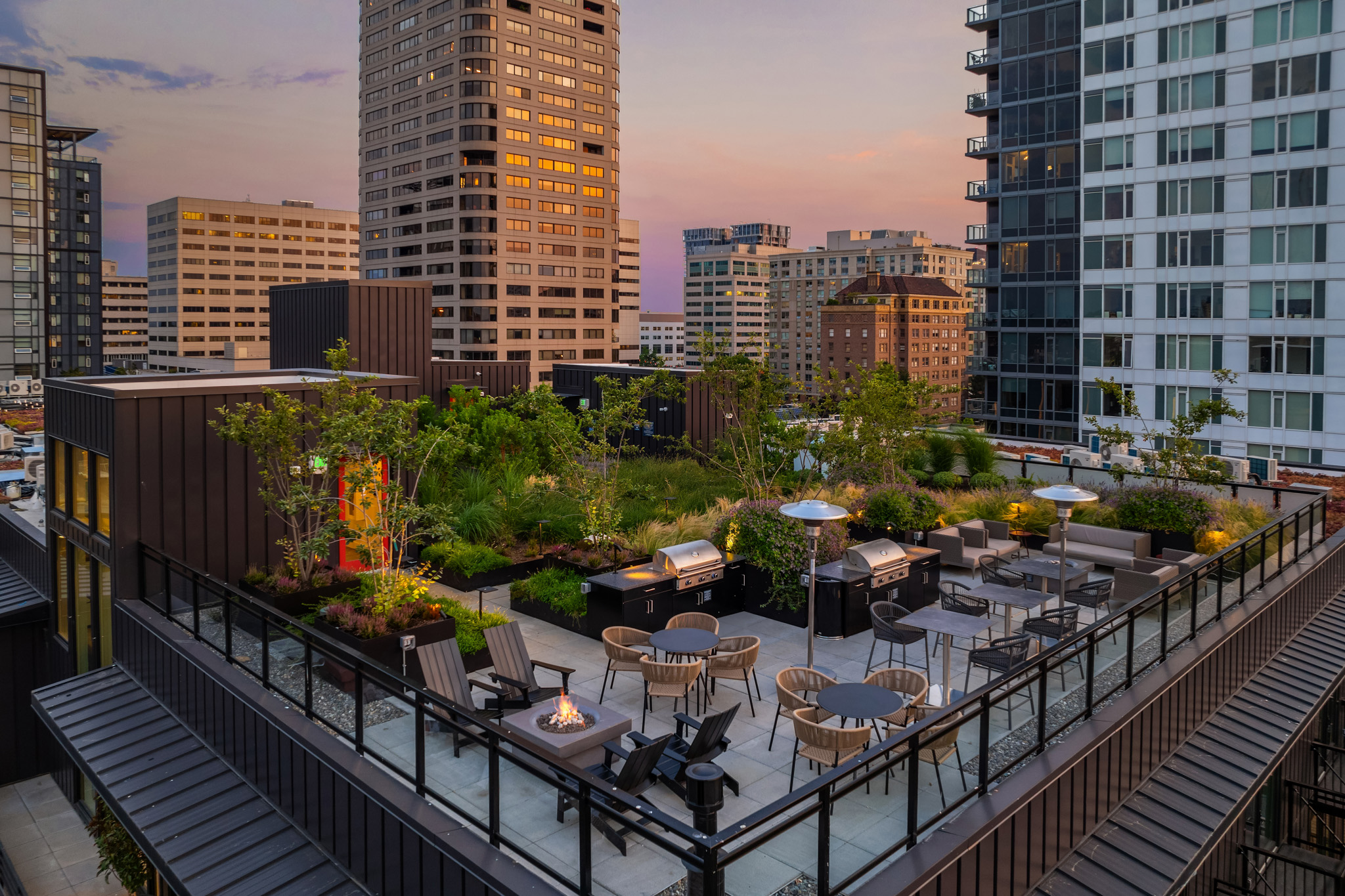 Rooftop garden terrace with modern seating, fire pit, and grilling area, showcasing lush greenery and city skyline views at twilight in Seattle.