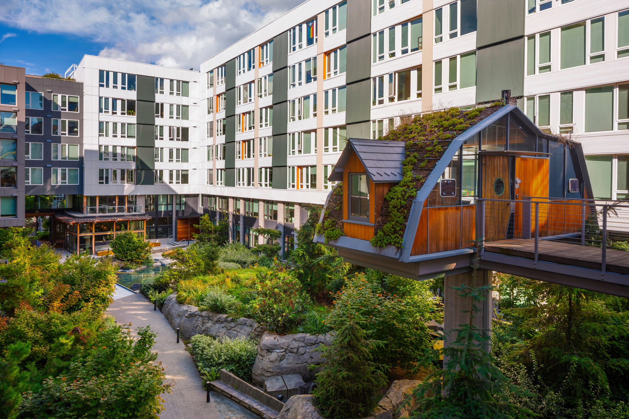 Modern apartment complex with a unique garden space and eco-friendly architecture, featuring a small wooden house with a green roof, nestled among lush landscaping in the Pacific Northwest.