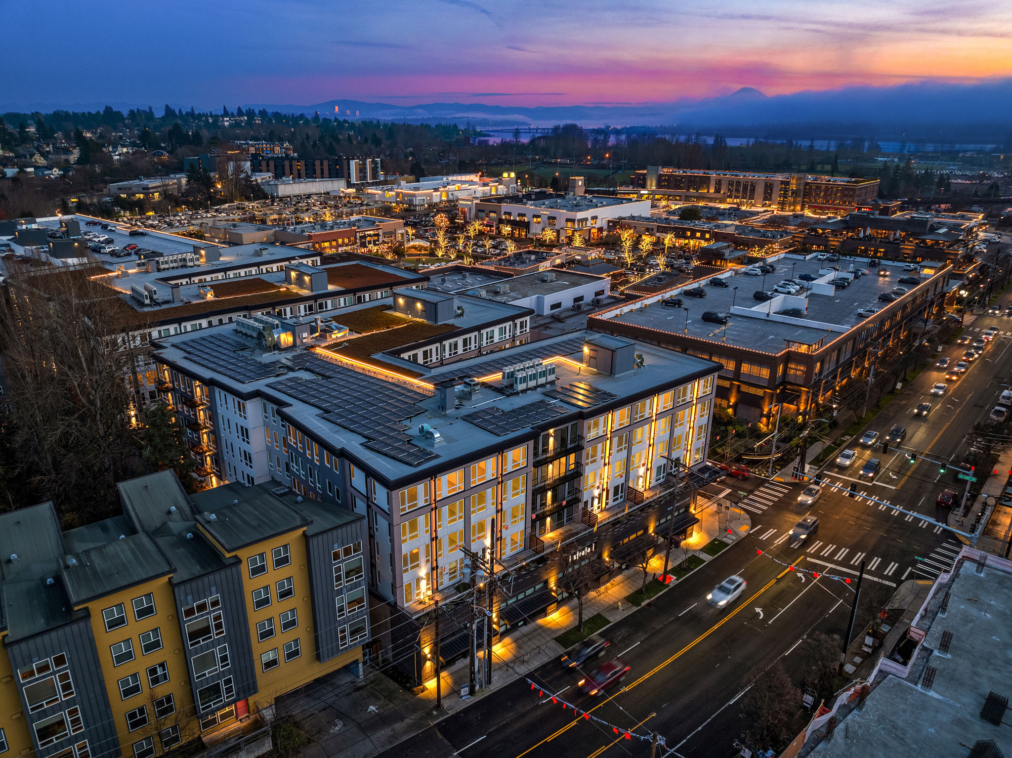 Aerial view of a modern mixed-use development at twilight in the Pacific Northwest, showcasing illuminated buildings, vibrant streets, and nearby mountains.