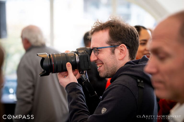 Candid shot of a photographer smiling while capturing images at a vibrant real estate event in Seattle, showcasing the lively atmosphere and engagement of attendees.