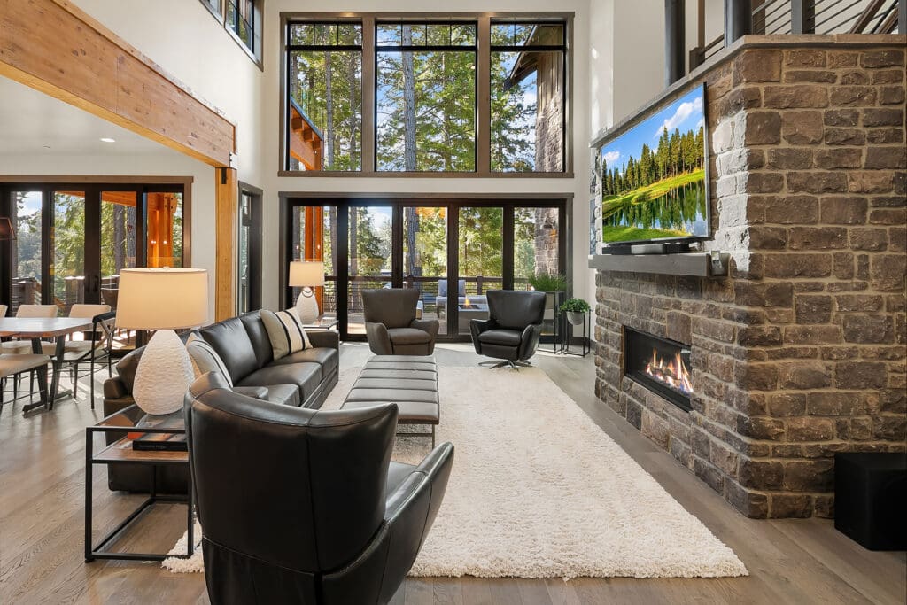 Modern living room with large windows showcasing a forest view, featuring a cozy fireplace, stylish black leather furniture, and warm wood accents, located in the Pacific Northwest.