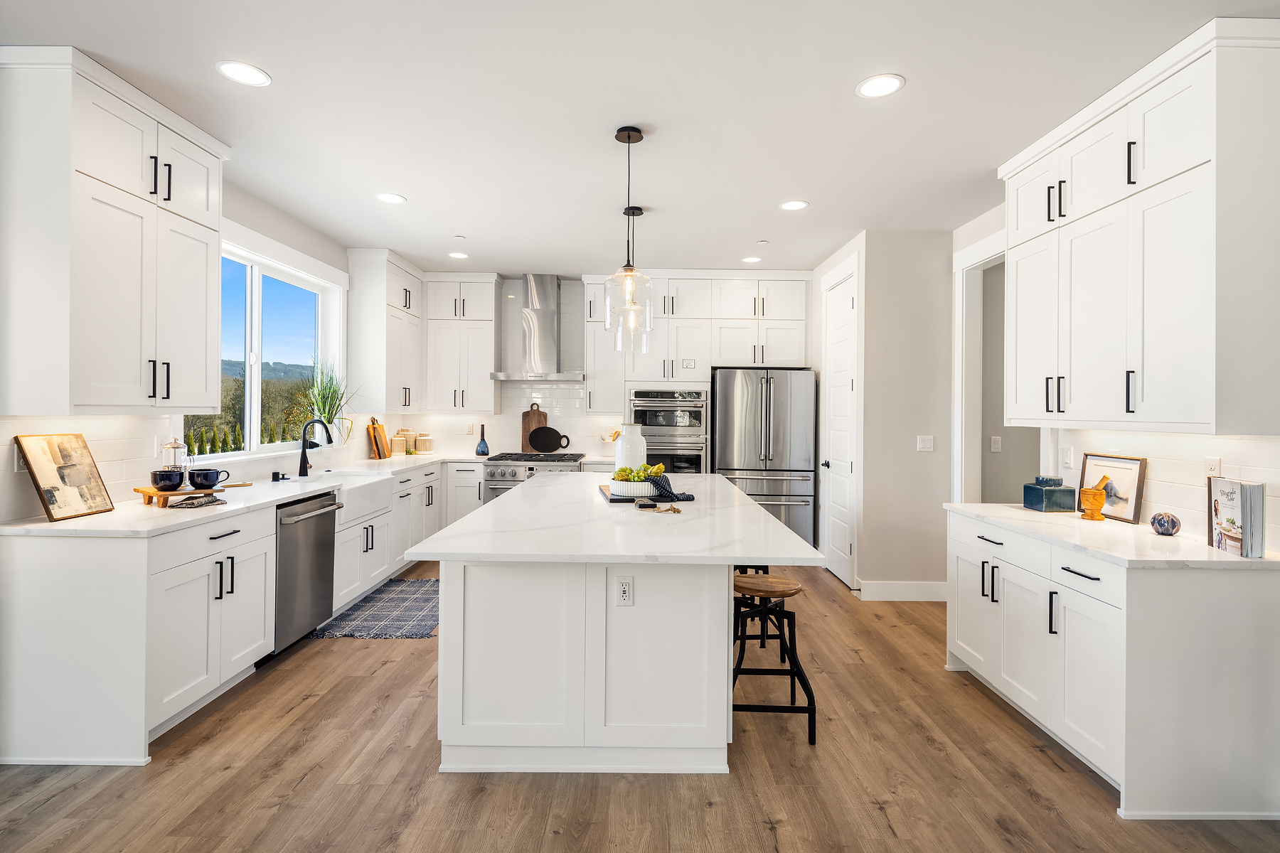 Modern kitchen featuring sleek white cabinetry, stainless steel appliances, and a spacious island with seating, illuminated by pendant lights. Bright and airy atmosphere with large windows showcasing scenic views, located in the Pacific Northwest.