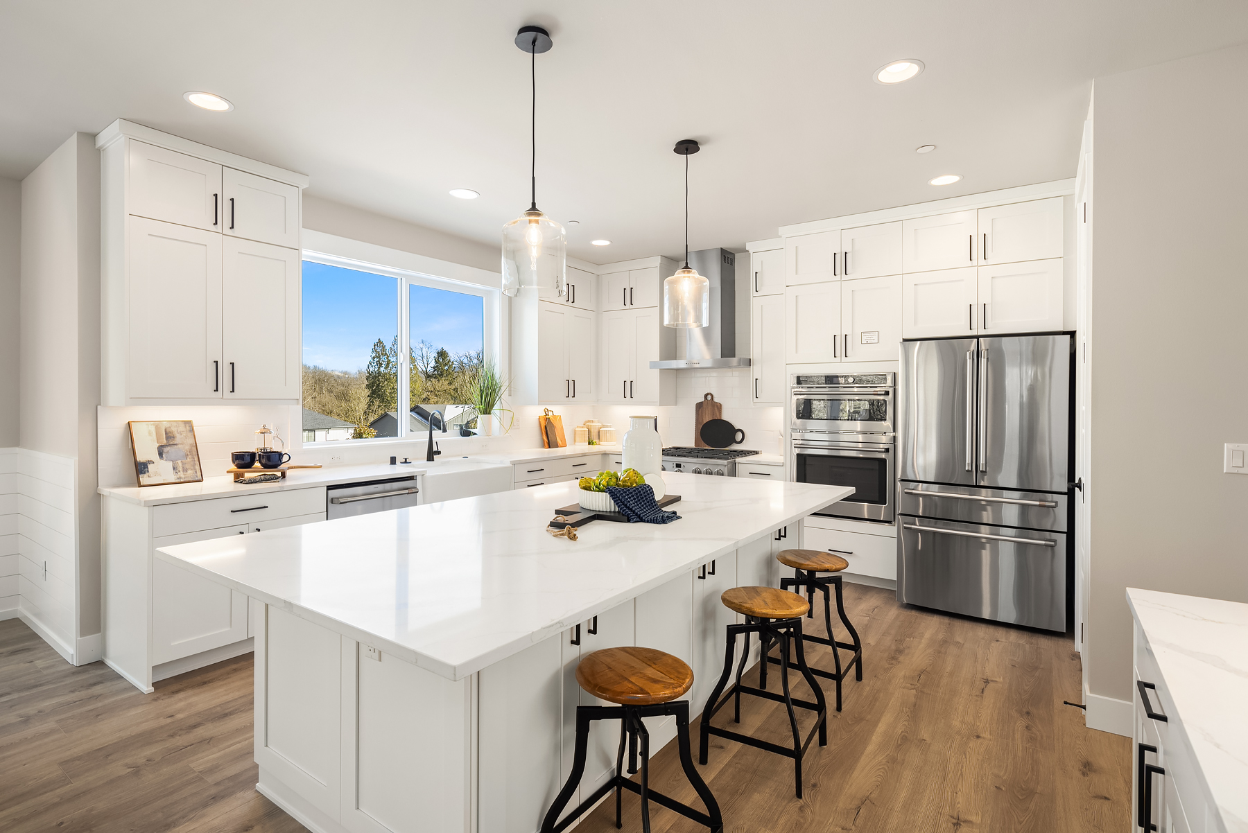 Modern kitchen with white cabinetry, stainless steel appliances, and a large island featuring a quartz countertop, located in the Pacific Northwest. Bright natural light floods the space through a large window, enhancing its inviting atmosphere.