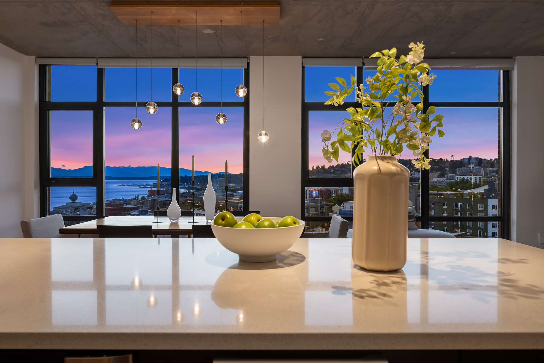 Modern kitchen with a sleek countertop, elegant decor, and stunning sunset views over Seattle's waterfront, featuring green apples and stylish vases.